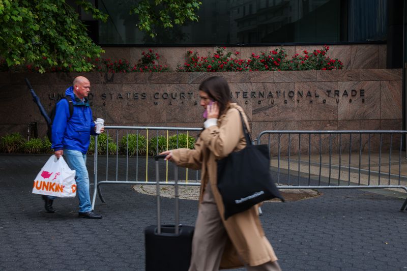Two individuals walking near the United States Court International Trade building, one carrying a bag and coffee, the other pulling a suitcase