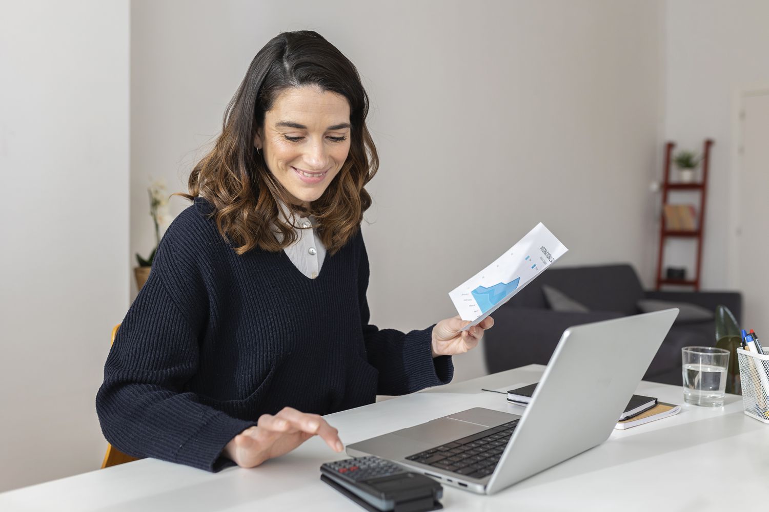 A happy woman uses a calculator and laptop at a desk.