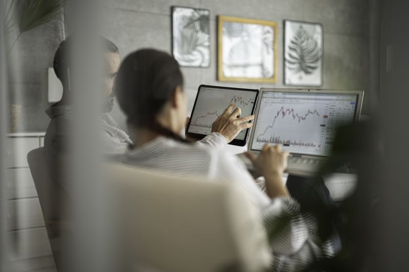 Two people sitting infront of computer monitors showing stock charts and having a discussion.