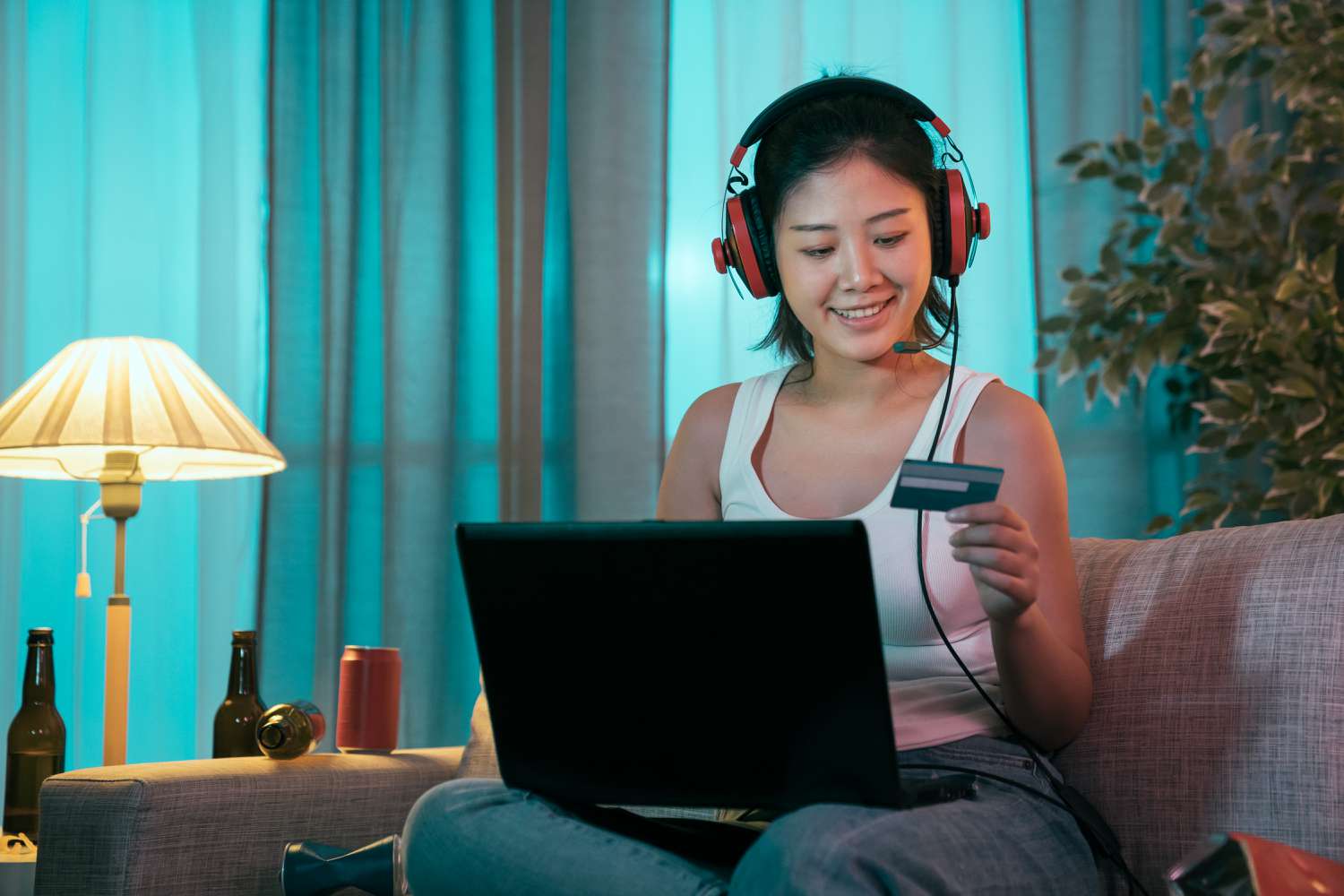 A happy woman wearing headphones sits on a couch with a laptop, smiling at a debit card in her hand.