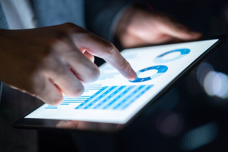 A close-up of a person's hand tapping a tablet screen displaying financial charts and tables.