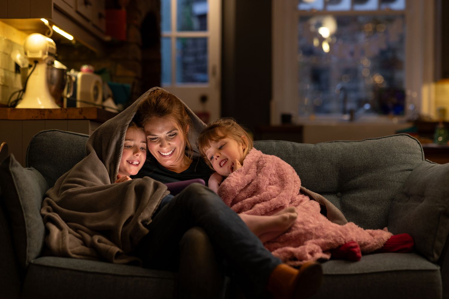 Mom and children sitting on the couch with blanket over their heads, enjoying a cosy night in the house that they are owner-occupants.