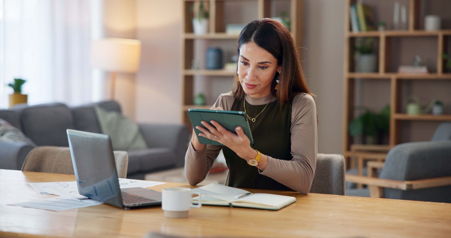 Woman sitting at a desk in her home office holding a tablet with a laptop on the desk.