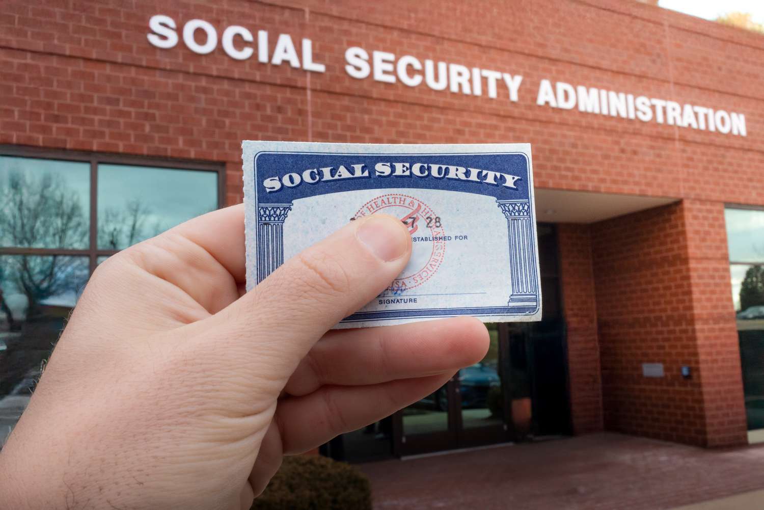 Someone holds in their hand a Social Security card with the building for the Social Security Administration in the background.