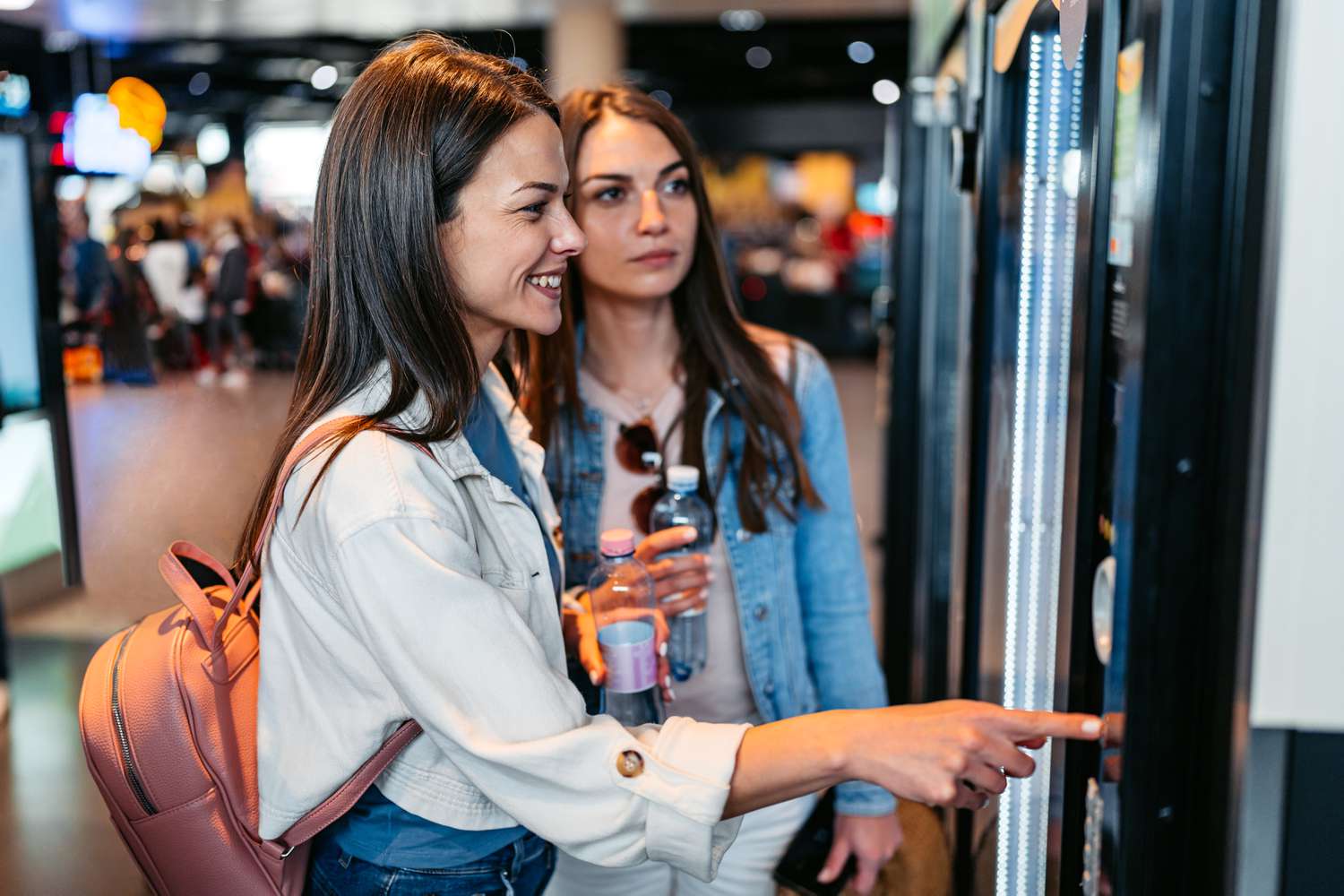 Two women holding water bottles stand in front of a vending machine at an airport in Austria.