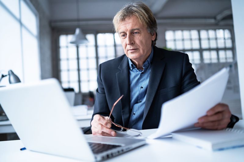 Businessman working at a desk looking at a laptop with papers in his hand