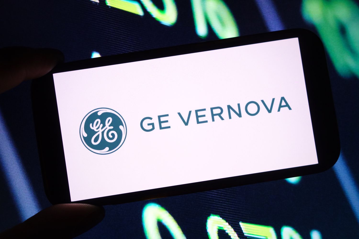 The blue-green GE Vernova is seen on a phone screen, in front of a screen with green numbers in the background.