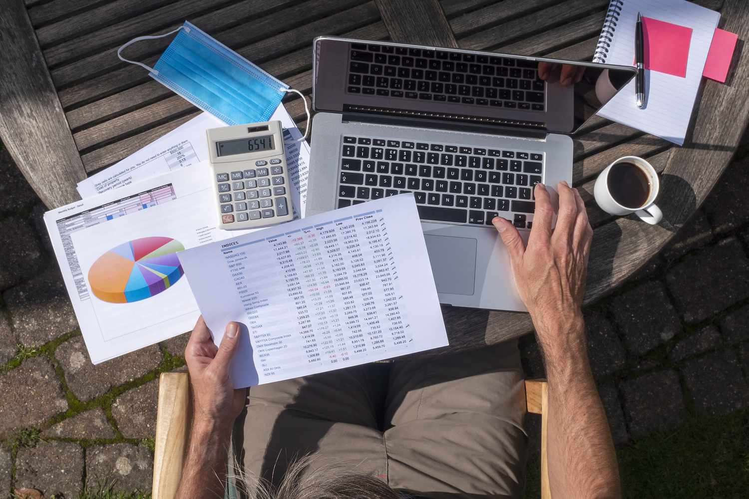 A person seated outside, picture taken from above looking down, as they review a list of stocks with one hand on a laptop.