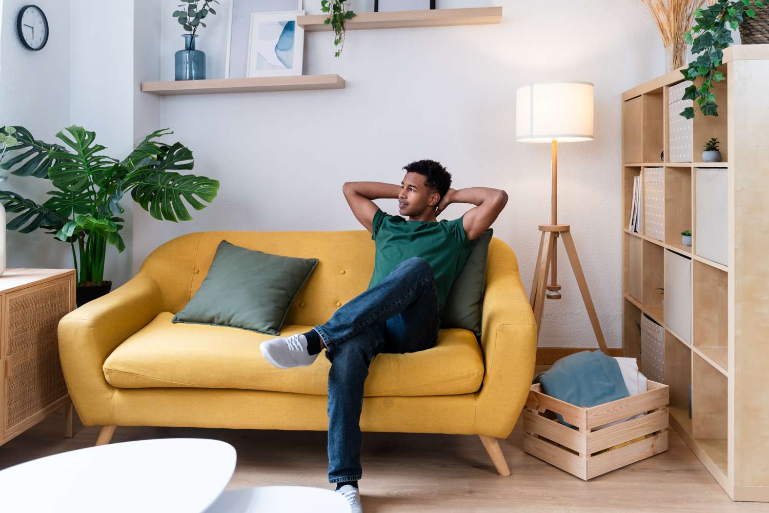 A man in jeans and a green shirt sits on a yellow couch in an apartment