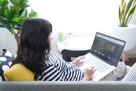 A woman sits on a couch with a laptop in her lap, looking at financial data on the computer.