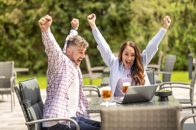 A couple sitting outside at a restaurant are looking at a laptop and excited about what's on it.