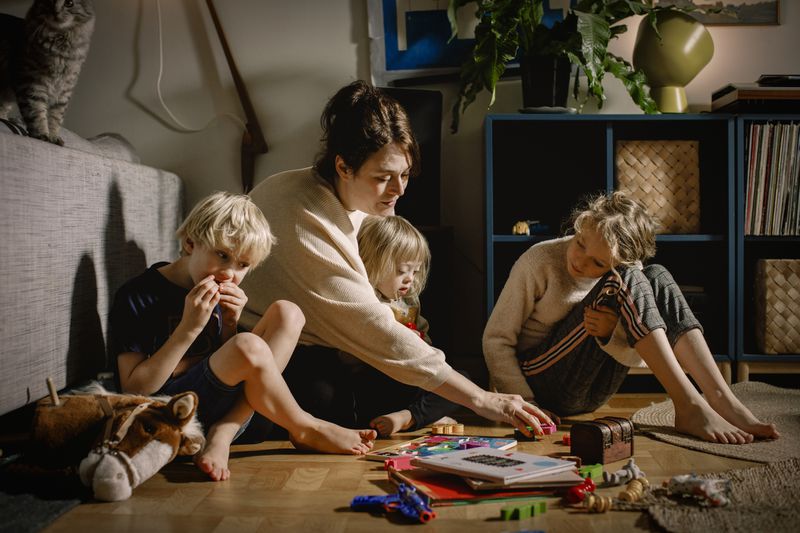 A mom sits on the floor with three kids, surrounded by toys and books.