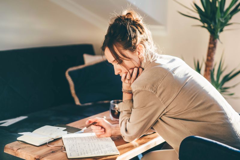A woman leans over a table in a living room, writing in a journal. 