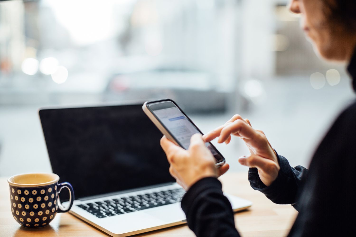 A person sitting at a desk with a laptop and mug in front of them. They're holding a smartphone in one hand.