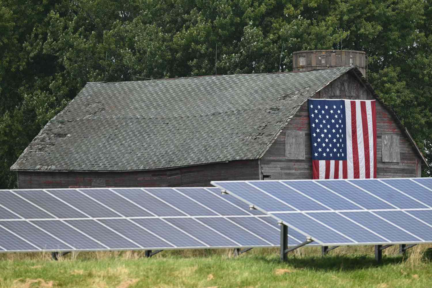 A grow of solar panels are seen in front of a barn with an American flag.