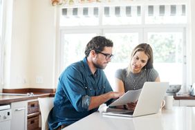 A man and a woman sit at the kitchen counter looking at papers and a laptop