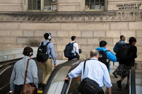 People on an escalator wear headphones and work bags outside of an ornate brick building.