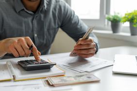 Man using calculator while holding money at desk with files, a computer, and an open notebook