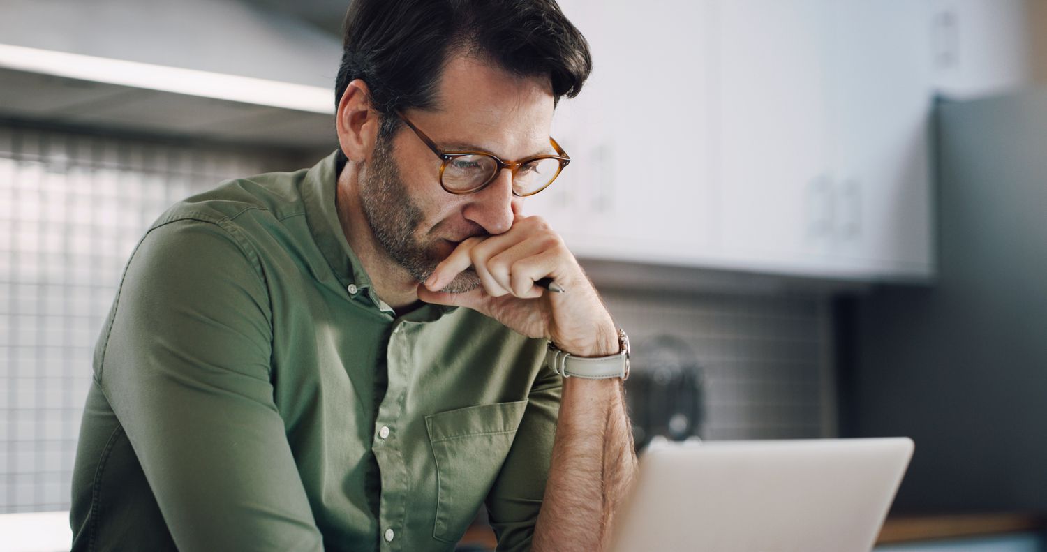 Man in his 40s sitting in his kitchen and seriously pondering financial information on his laptop