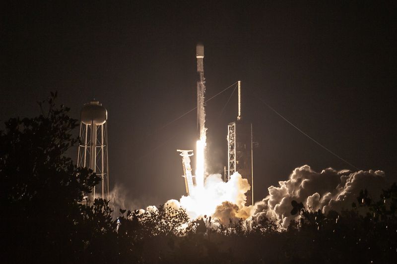 Fire and smoke is seen below a launching SpaceX rocket, with a dark sky in the background.