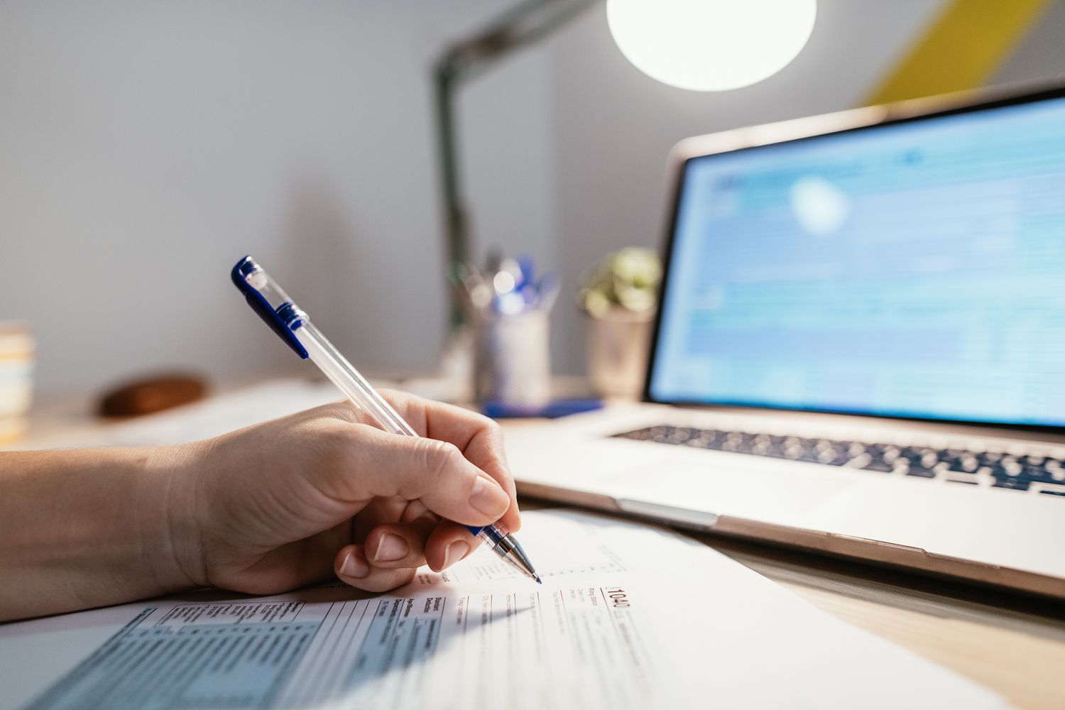 Close up of a hand holding a pen filling out tax forms with a laptop in the background