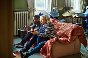 Two young adults playing console video games sitting on a couch in a small apartment.
