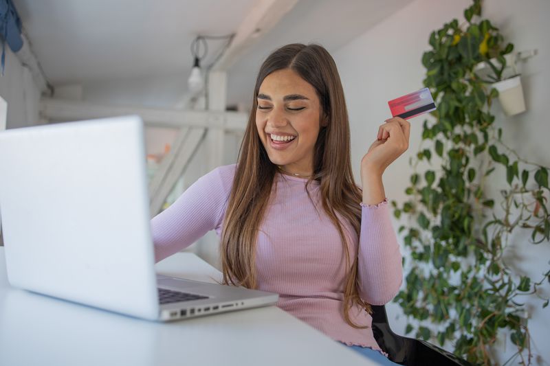 A woman smiles happily at her computer while holding up a debit card.