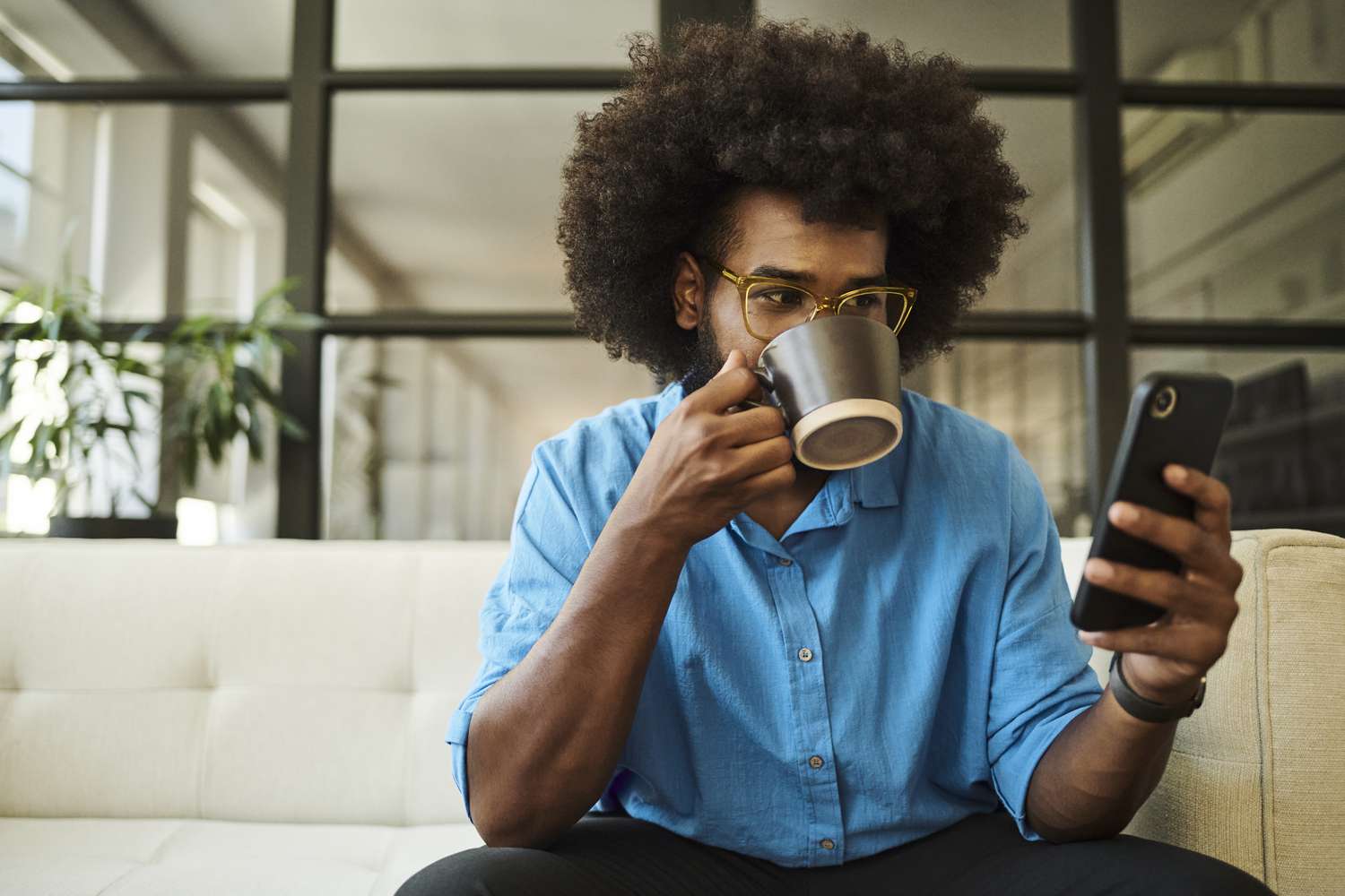 A man in a blue short and glasses drinks from a mug and stares at a smartphone