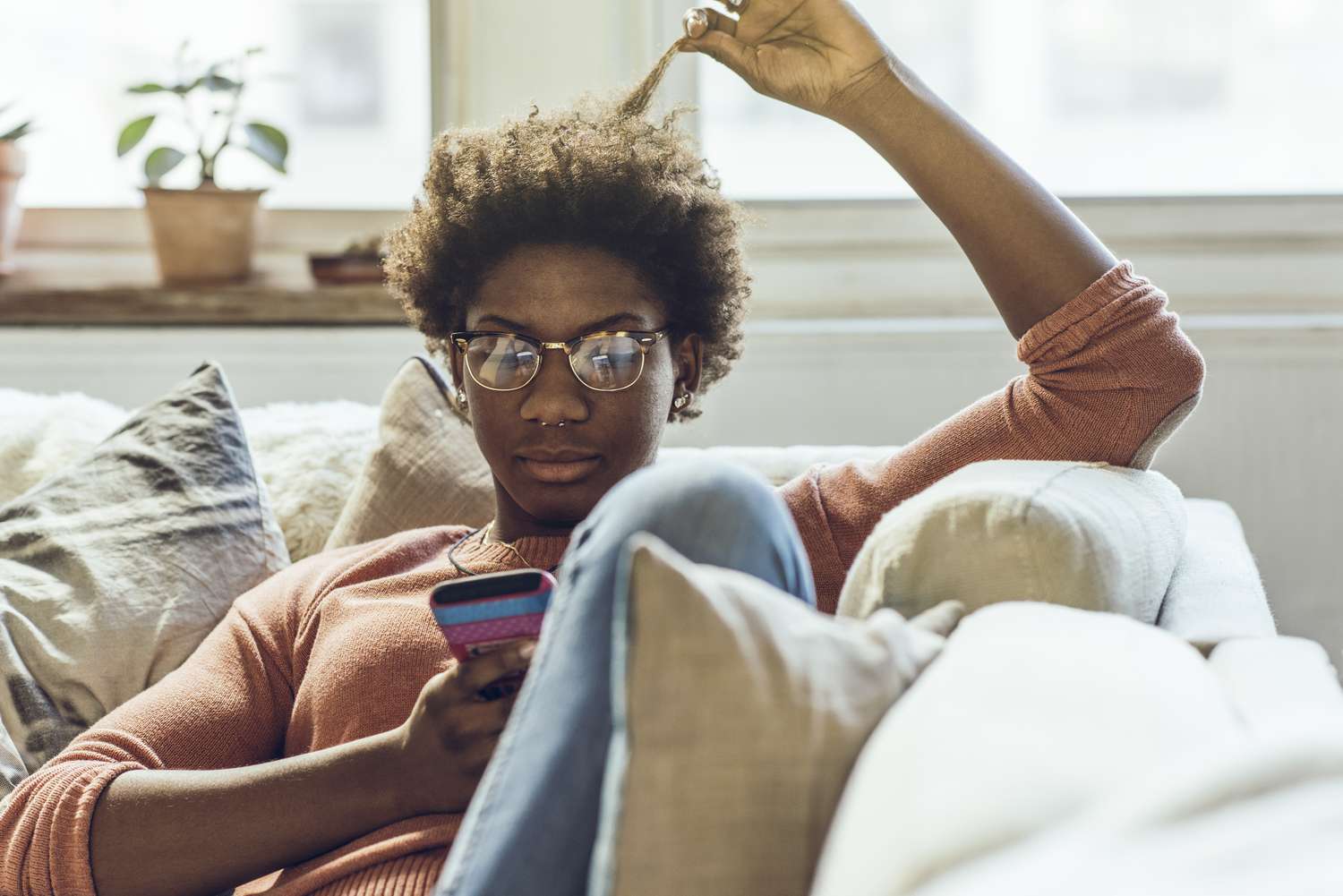 A woman with glasses sits on a couch reading something on her smartphone