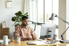 Black male wearing glasses sits at work desk in front of desktop computer