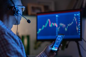 active investor at a computer screen, holding a phone, both showing stock charts. The trader is wearin a headset and sitting in the dark. 