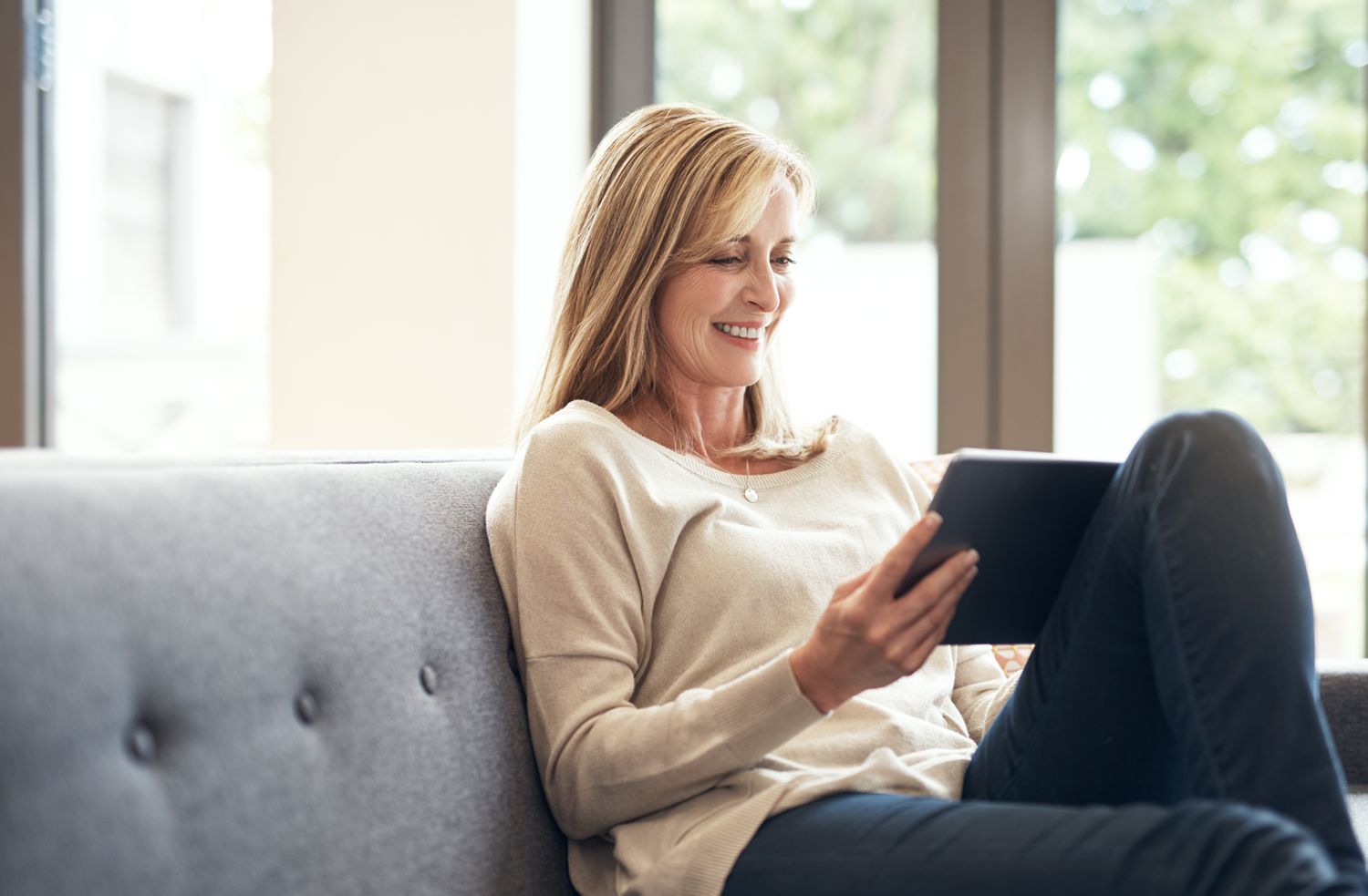 Woman in her 40s sitting on her sofa and looking happily at bank rates on her tablet