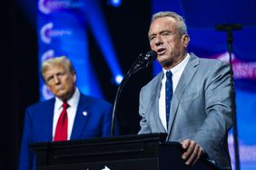 Robert F. Kennedy Jr. speaks at a podium while Donald Trump stands behind him in front of a blue background.