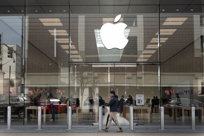 People walk past the glass facade of an Apple store, with the Apple logo hanging above them.
