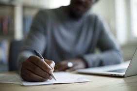 Person at a laptop writing on paper with a pen