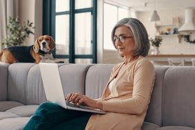 An older woman sits on a couch typing on a laptop while her dog lounges on the couch next to her