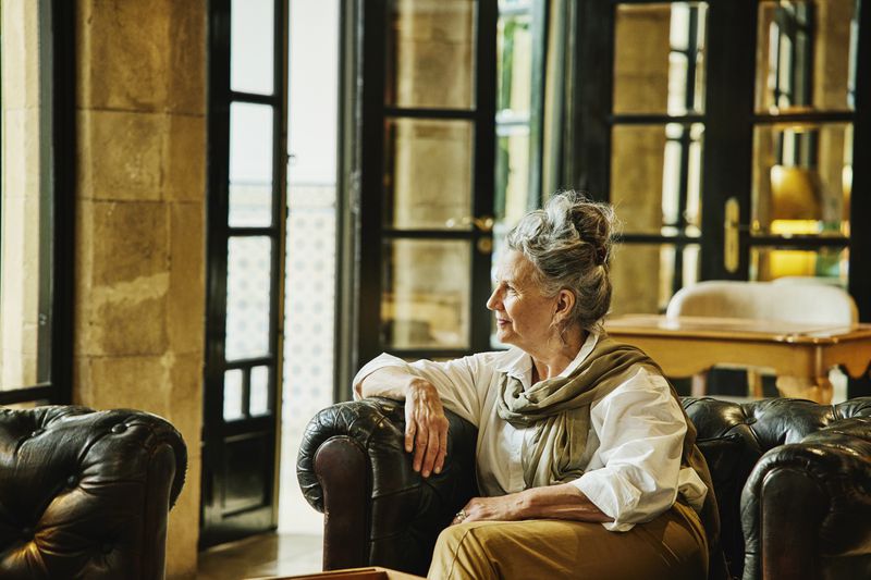 Older adult seated in a room with large windows, looking contemplative