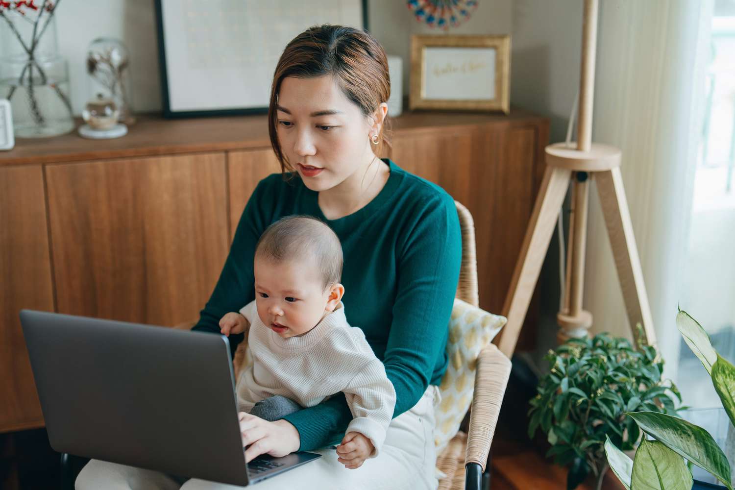 Woman holding a baby on her laptop while she works on a laptop at home. 
