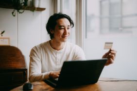 A man sits a table with a laptop, holding a credit card in his hand and smiling.