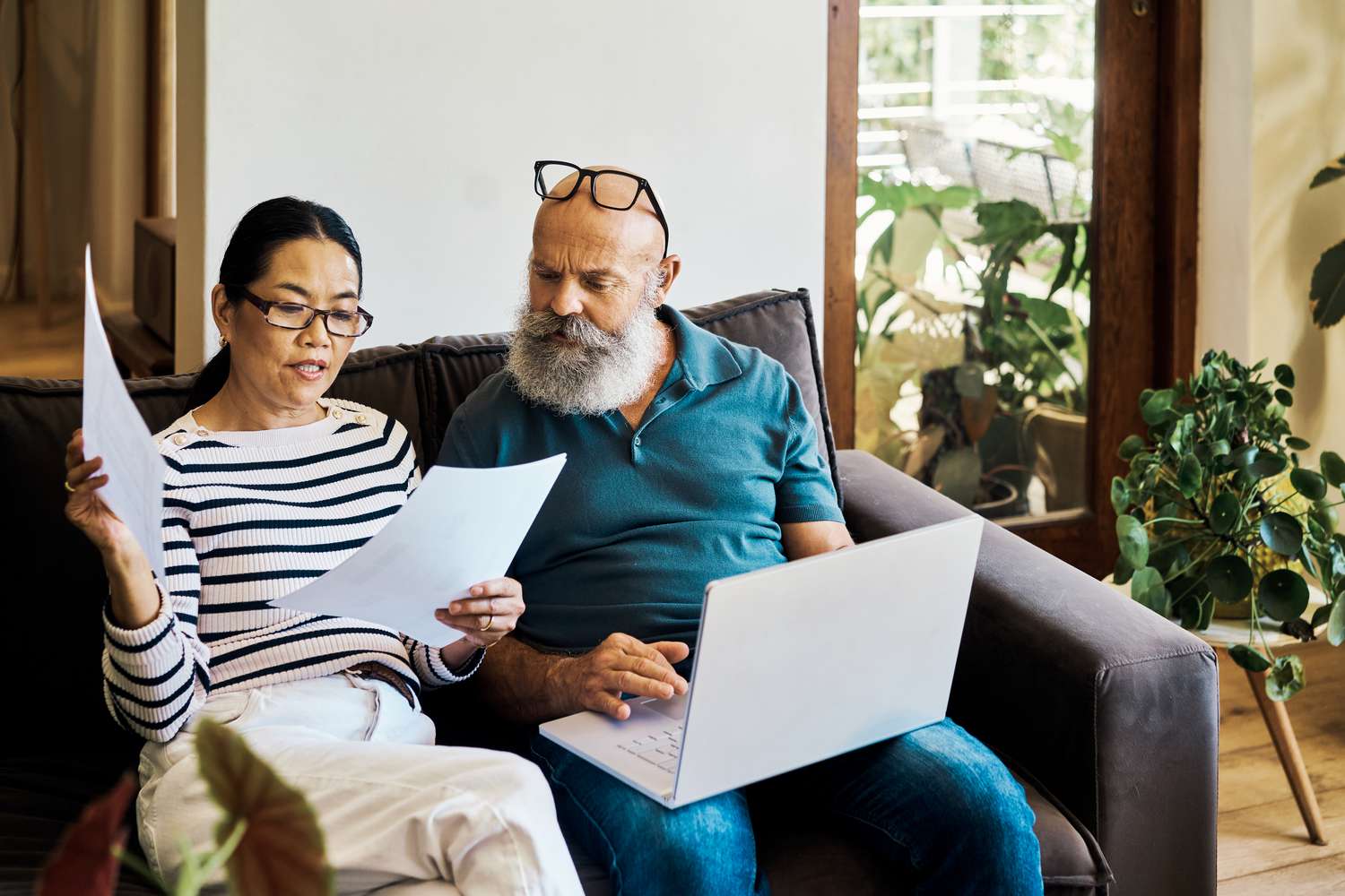 An adult couple sit on a sofa with one holding paperwork and the other using a laptop and review their retirement plan activity.