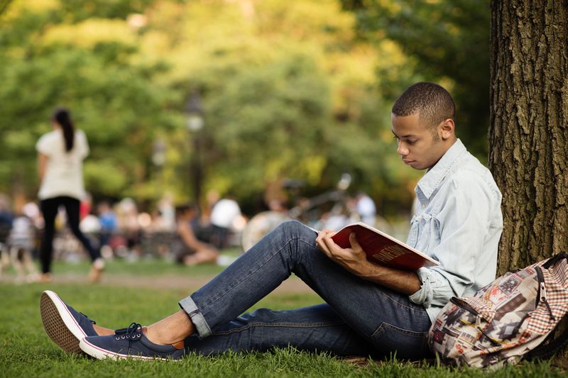 Side view of man studying while sitting by tree trunk in campus