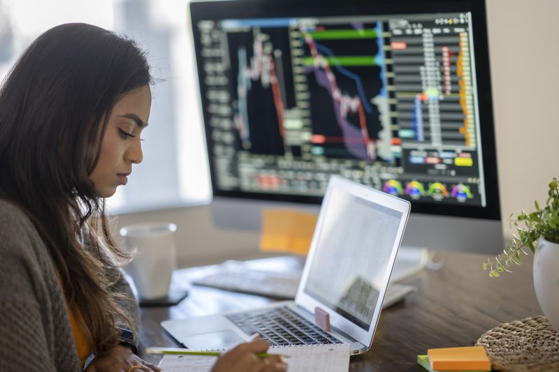 Woman working on a laptop with a larg monitor on her desk displaying charts and other data.