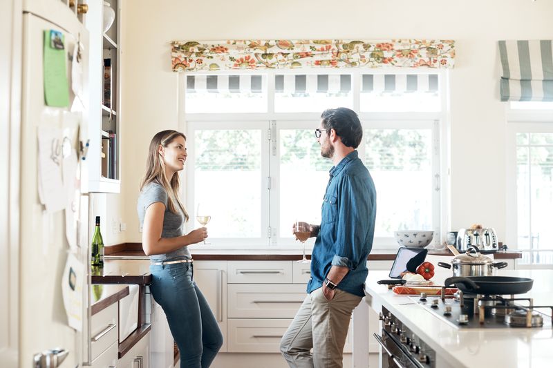 A man and a woman stand in a kitchen talking while holding glasses of wine