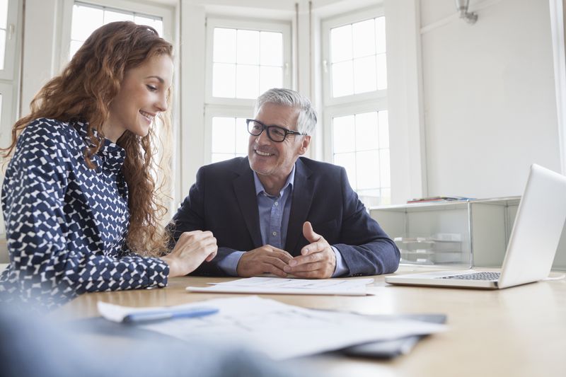 Two executives talk at a desk with documents.