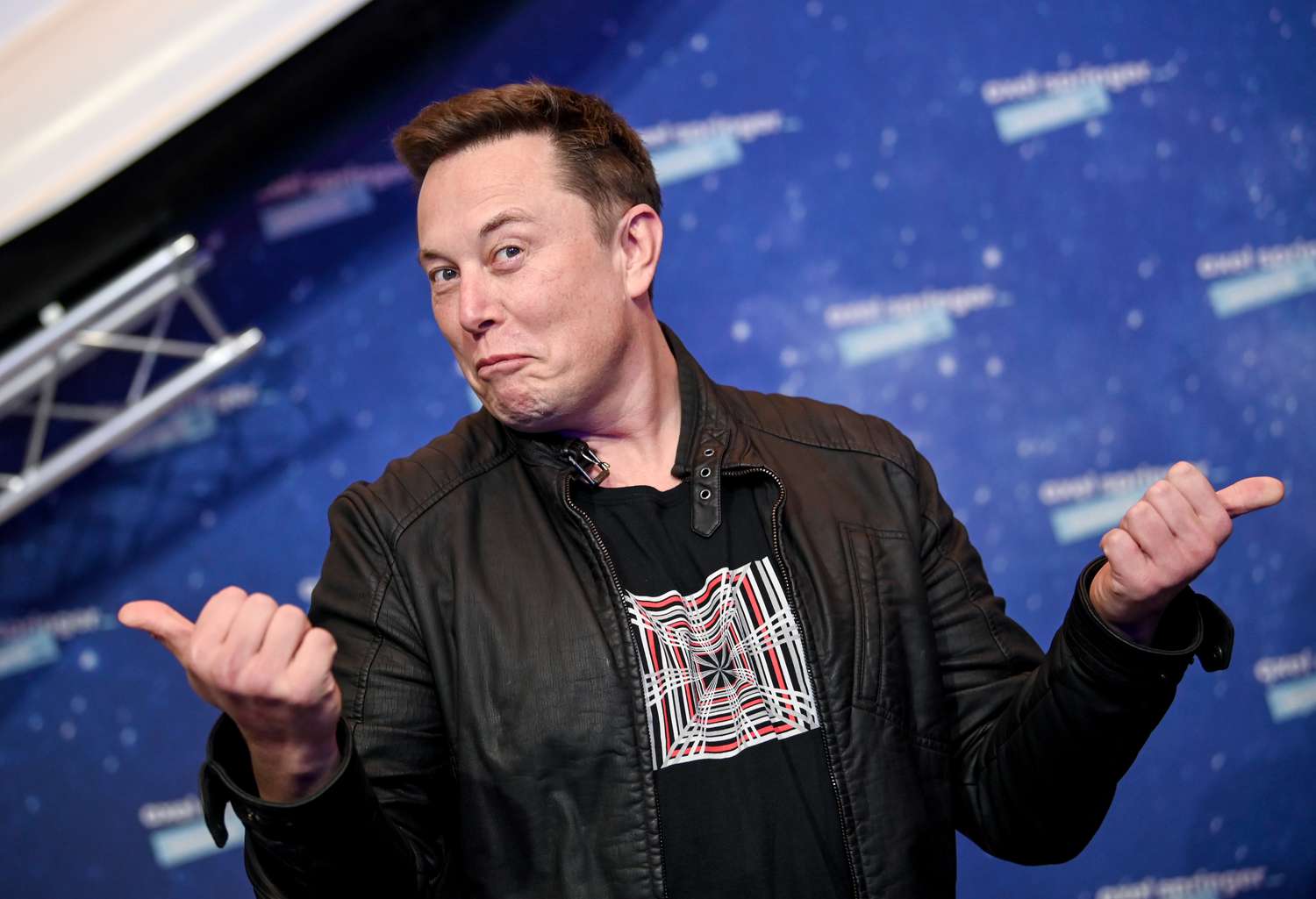 Elon Musk shrugging while giving two thumbs up on a red carpet. 