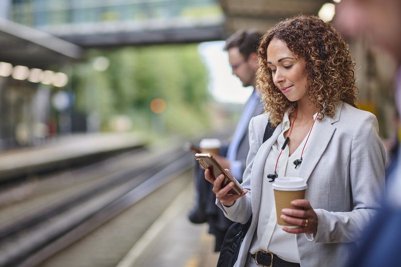Woman on a train platfor reading her phone while holding a cup of coffee. 