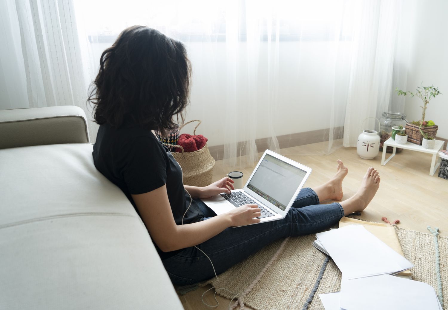 A woman in a black shirt and jeans sits barefoot on the floor with a laptop and paperwork