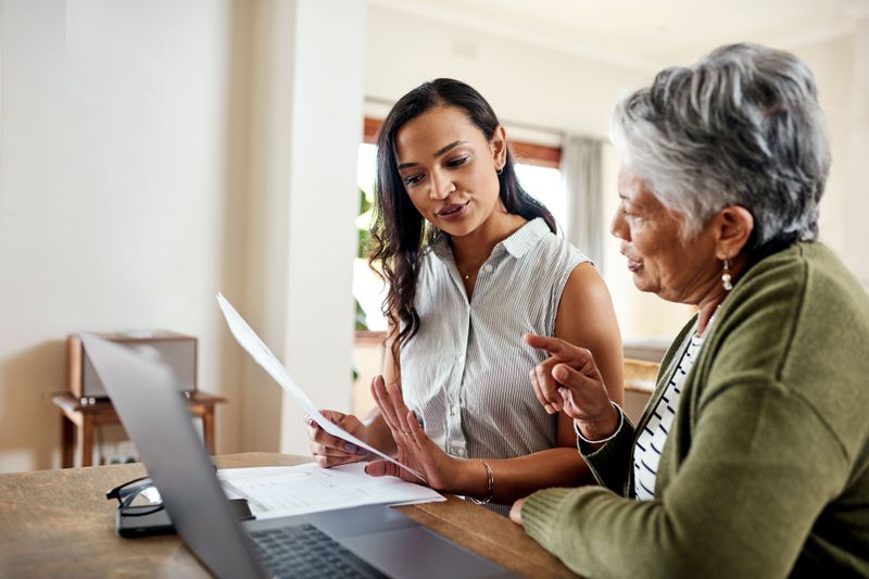 Two women discussing paperwork in front of a laptop at home