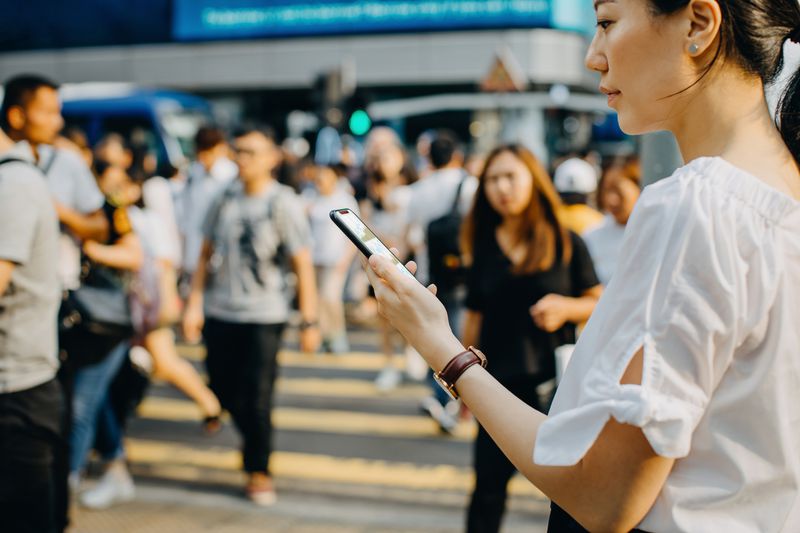 Young Asian woman checking financial trading data on smartphone in downtown city street, against busy pedestrian walkway and city traffic