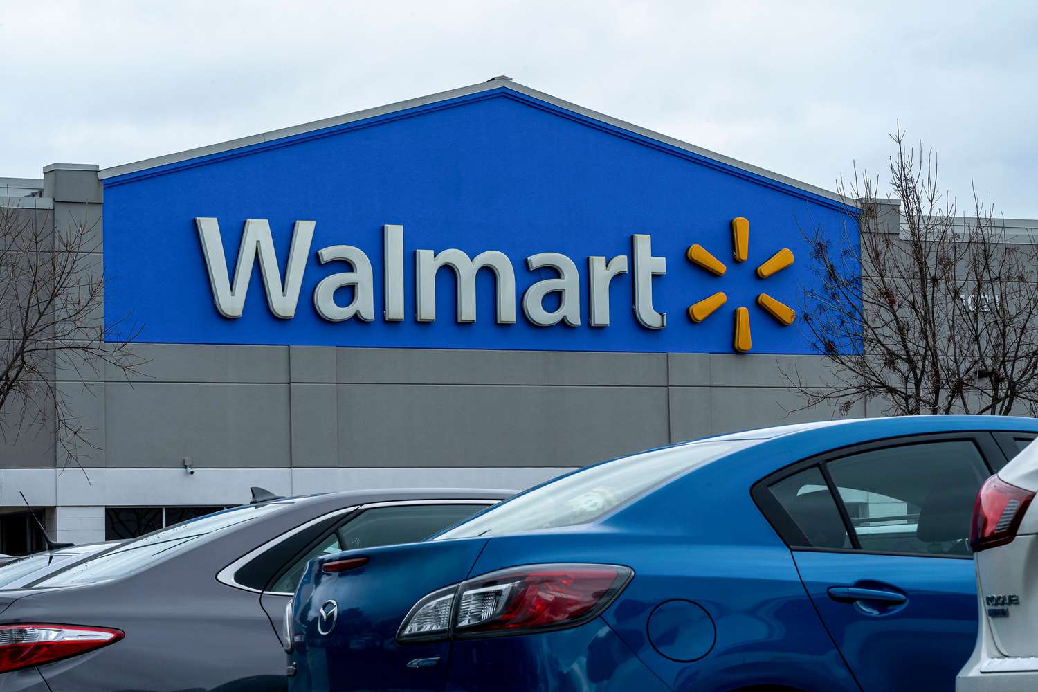 The Walmart logo is seen on the blue front of a store with several cars in the foreground.